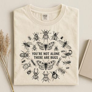 Op de afbeelding: Cr&egrave;mekleurig T-shirt met een cirkelvormig ontwerp van zwarte insectenillustraties, waaronder vlinders, kevers en een schorpioen. De tekst "YOU'RE NOT ALONE THERE ARE BUGS" staat in het midden. Het shirt heeft een Comfort Colors-label.