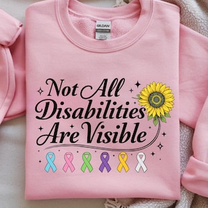 May include: Pale pink crew neck sweatshirt featuring the message "Not All Disabilities Are Visible" in black script. A sunflower graphic and awareness ribbons in various colours are also displayed. The sweatshirt is a casual style.