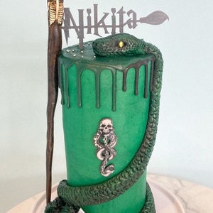 Könnte beinhalten: Ein grüner Kuchen, verziert mit einer Schlange, einem Schädel und grünem, tropfendem Zuckerguss. Der Kuchen ist mit einem schwarz-weißen Schild mit der Aufschrift "Nikita" gekrönt.