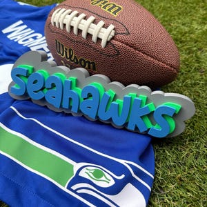 Può includere: La parola "Seahawks" in 3D blu e verde con un pallone da football marrone e bianco in cima. Il pallone da football ha la scritta "Wilson" su di esso.