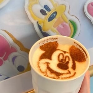 May include: A white ceramic mug with a Mickey Mouse design in cinnamon on top of the latte. The mug is being held by a hand with blue nail polish.
