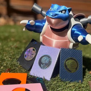May include: A blue and pink 3D-printed Blastoise figure is positioned above several trading card holders. The holders are in various colours, including black, purple, and blue, each with a circular design featuring a Pokémon character.