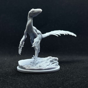 Może przedstawiać: Szara, trójwymiarowa miniatura dinozaura, stojąca na szarej podstawie. Dinozaur ma długi ogon i pióra.