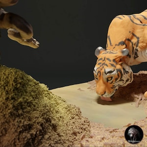 Pode incluir: Um close-up de uma figura de tigre bebendo de uma pequena poça de água. O tigre é feito de argila laranja, preta e branca e tem detalhes realistas. A figura está sentada em uma superfície arenosa marrom e verde.