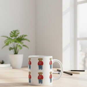 Op de afbeelding: Witte keramische koffiemok met een herhalend patroon van een teddybeer met een rode trui met "USA" erop, een spijkerbroek en sneakers. De mok heeft een wit handvat en staat op een lichtgekleurd houten oppervlak.