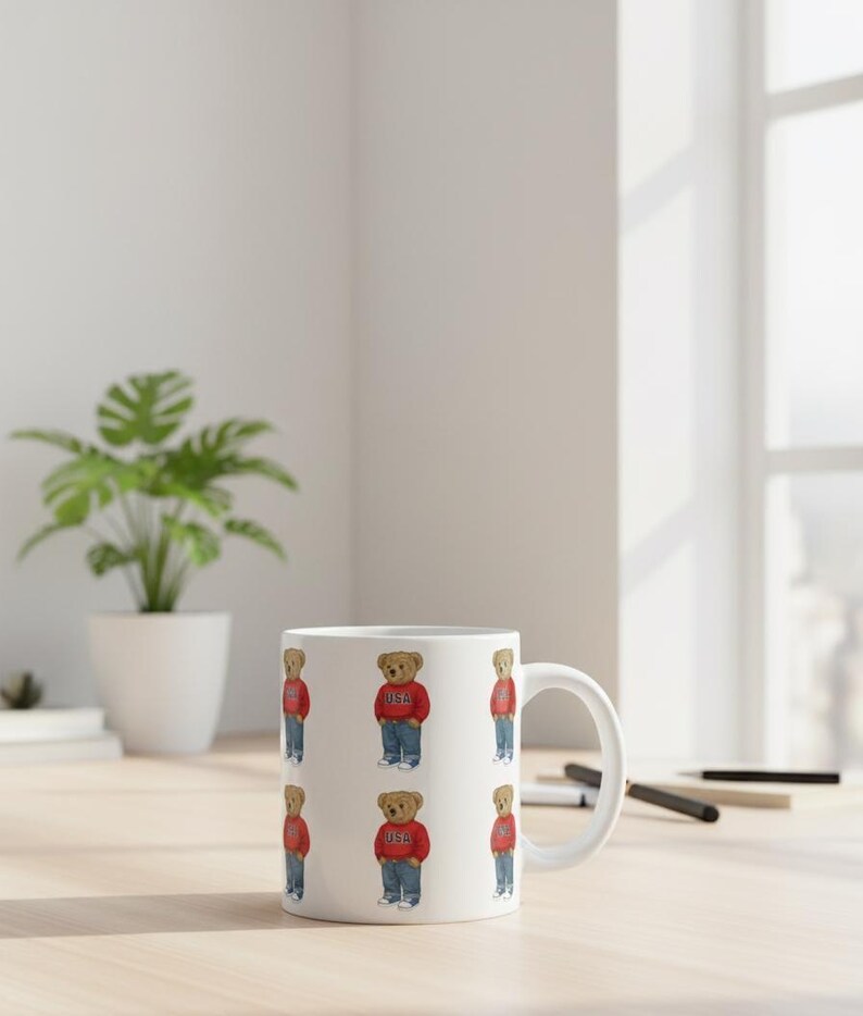 Op de afbeelding: Witte keramische koffiemok met een herhalend patroon van een teddybeer met een rode trui met "USA" erop, een spijkerbroek en sneakers. De mok heeft een wit handvat en staat op een lichtgekleurd houten oppervlak.