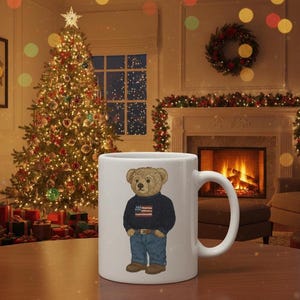Op de afbeelding: Witte keramische mok met een cartoon teddybeer met een marineblauwe trui met een Amerikaans vlagontwerp en een blauwe jeans. De mok staat tegen een feestelijke kerstsc&egrave;ne.