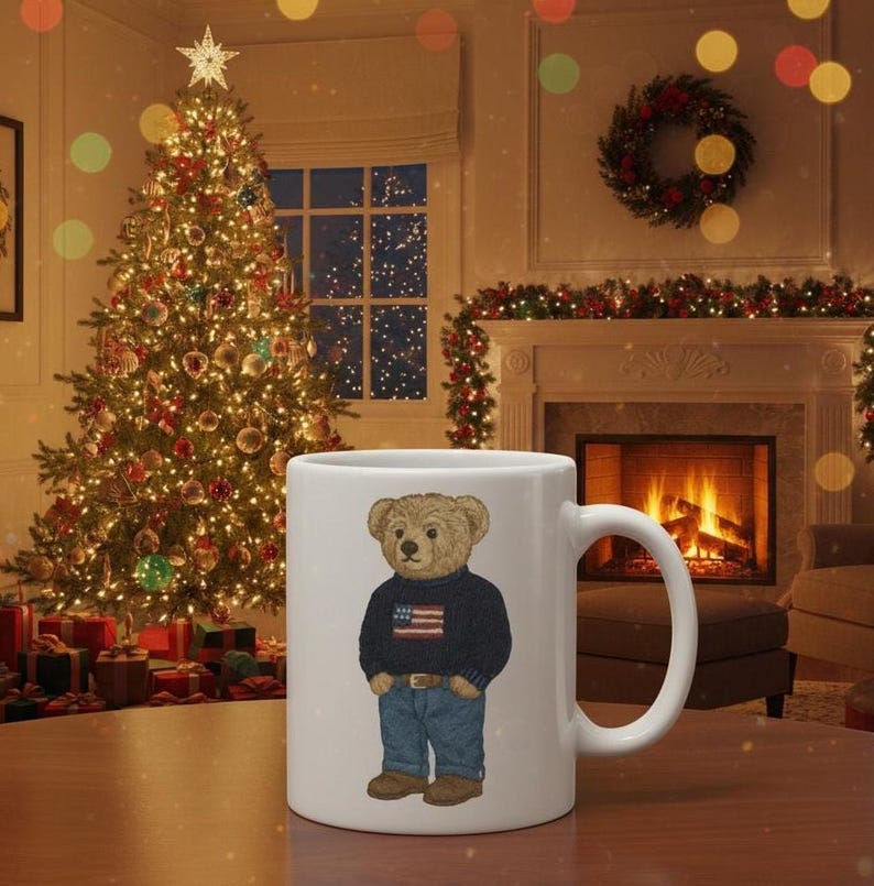 Op de afbeelding: Witte keramische mok met een cartoon teddybeer met een marineblauwe trui met een Amerikaans vlagontwerp en een blauwe jeans. De mok staat tegen een feestelijke kerstsc&egrave;ne.