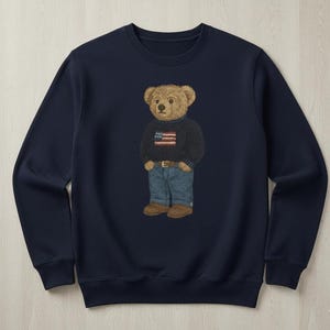 Op de afbeelding: Marineblauw sweatshirt met een geborduurde teddybeer die een trui draagt met een Amerikaans vlagontwerp, een blauwe spijkerbroek en bruine schoenen. De beer is de centrale focus, met een gedetailleerde, realistische stijl. De sweatshirt heeft een ronde hals.