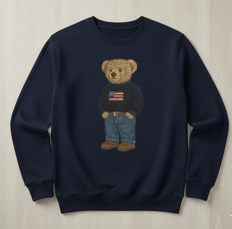 Op de afbeelding: Marineblauw sweatshirt met een geborduurde teddybeer die een trui draagt met een Amerikaans vlagontwerp, een blauwe spijkerbroek en bruine schoenen. De beer is de centrale focus, met een gedetailleerde, realistische stijl. De sweatshirt heeft een ronde hals.