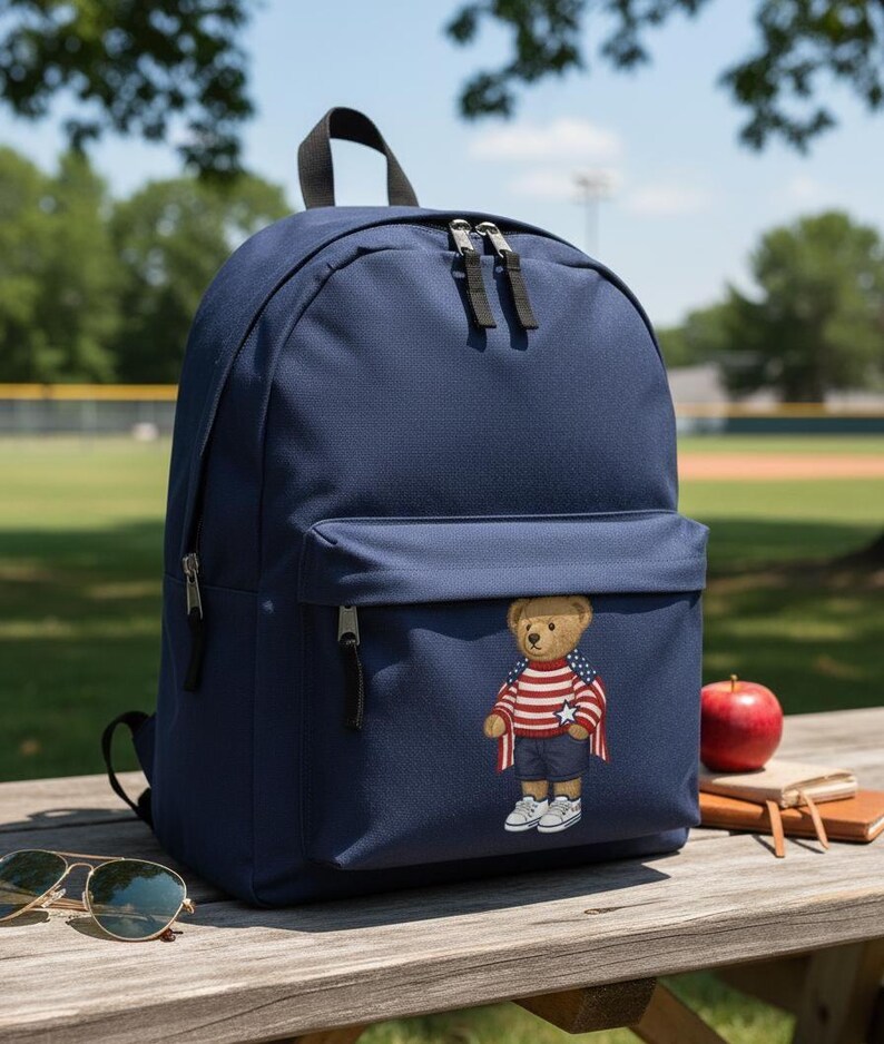 Op de afbeelding: Marineblauwe rugzak met een voorvak met een teddybeer in een patriottische outfit. De rugzak heeft een handvat aan de bovenkant, twee compartimenten met ritssluiting en verstelbare schouderbanden. Een zonnebril en een rode appel liggen op een houten oppervlak.