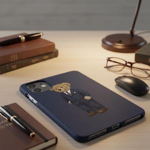 Op de afbeelding: Een marineblauwe telefoonhoes met een cartoonbeer in pak en stropdas. De hoes is omgeven door een pen, notitieboek, boeken, bril, een computermuis en een bureaulamp. De hoes is ontworpen voor een smartphone.