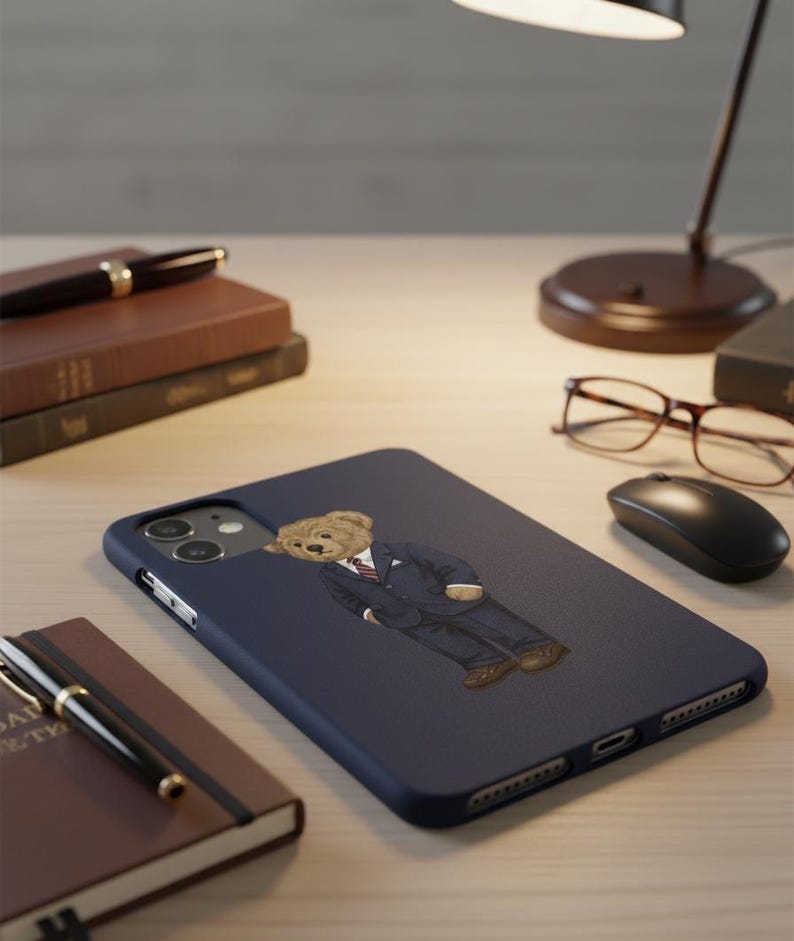 Op de afbeelding: Een marineblauwe telefoonhoes met een cartoonbeer in pak en stropdas. De hoes is omgeven door een pen, notitieboek, boeken, bril, een computermuis en een bureaulamp. De hoes is ontworpen voor een smartphone.