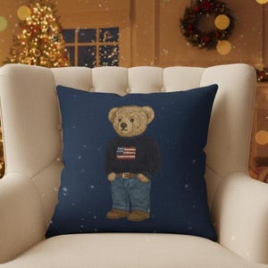Op de afbeelding: Marineblauw decoratief kussen met een teddybeer met een trui met een Amerikaanse vlag, een blauwe spijkerbroek en bruine laarzen. Het kussen ligt op een witte fauteuil voor een kerstboom.