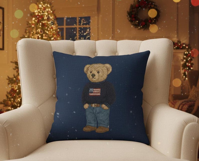 Op de afbeelding: Marineblauw decoratief kussen met een teddybeer met een trui met een Amerikaanse vlag, een blauwe spijkerbroek en bruine laarzen. Het kussen ligt op een witte fauteuil voor een kerstboom.