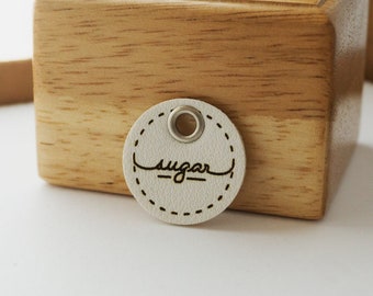 May include: A white round leather tag with a stitched edge and a metal eyelet. The tag has the word "sugar" written in cursive on it.