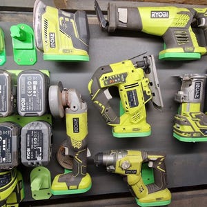 Op de afbeelding: Een verzameling Ryobi elektrisch gereedschap in geel, grijs en groen, waaronder een reciprozaag, decoupeerzaag, boor en slijper. Er zijn ook verschillende batterijpakketten en een oplader zichtbaar, allemaal gemonteerd op een houten oppervlak.