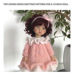 Puede incluir: Una muñeca con un vestido rosa tejido con ribete de encaje blanco, diseñado para una muñeca de 25,4 cm. La muñeca tiene cabello rizado oscuro, ojos azules y un lazo rosa. El texto dice "TOP DOWN DRESS KNITTING PATTERN FOR A 10 INCH DOLL."