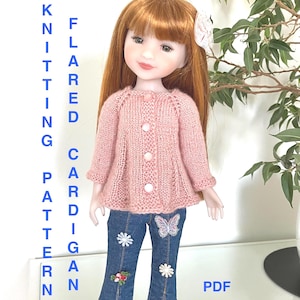 Puede incluir: Una muñeca con un cárdigan acampanado rosa tejido con tres botones, combinado con vaqueros azules con adornos florales y de mariposas. La muñeca tiene el pelo rojo y un accesorio blanco para el pelo. El texto en la imagen dice "Knitting Pattern Flared Cardigan PDF".