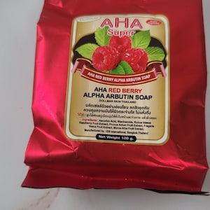 May include: A red foil package of AHA Red Berry Alpha Arbutin Soap. The label features a raspberry illustration with green leaves. The text on the label includes the product name, ingredients, and manufacturer information. Net weight is 100 g.