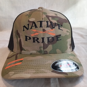 Könnte beinhalten: Eine Camouflage-Trucker-Kappe mit schwarzem Netzrücken. Die Kappe hat eine schwarze Stickerei mit der Aufschrift "NATIVE PRIDE" und zwei sich kreuzenden orangen Pfeilen in der Mitte.