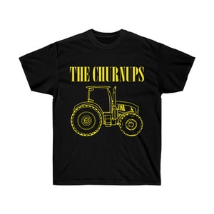 Puede incluir: Camiseta negra con un contorno amarillo de un tractor y el texto "THE CHURNUPS" encima. El dise&ntilde;o del tractor incluye ruedas grandes, una cabina y un tubo de escape. La camiseta es de un material suave.