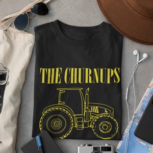 Puede incluir: Camiseta negra con el texto "THE CHURNUPS" en amarillo y un contorno amarillo de un tractor. La imagen incluye un sombrero marr&oacute;n, gafas de sol, una c&aacute;mara y otros accesorios.