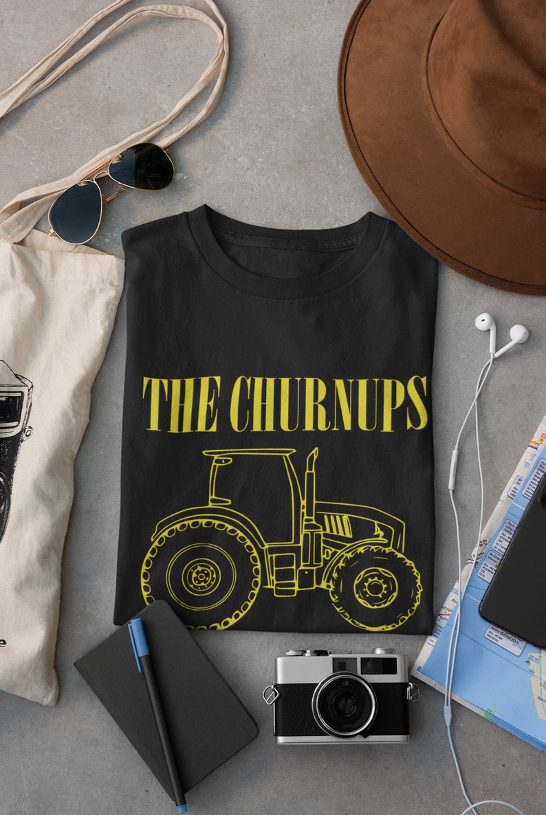 Puede incluir: Camiseta negra con el texto "THE CHURNUPS" en amarillo y un contorno amarillo de un tractor. La imagen incluye un sombrero marr&oacute;n, gafas de sol, una c&aacute;mara y otros accesorios.