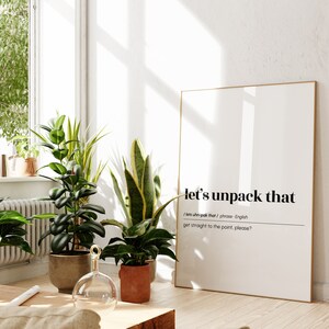 May include: A framed print with the text "let's unpack that" and a definition of the phrase. The print is hanging on a white wall in a room with plants.