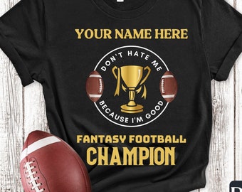 May include: Black t-shirt with gold text that reads "Your Name Here", "Don't Hate Me Because I'm Good", "Fantasy Football Champion". The text is surrounded by a circle with a football and a trophy inside.