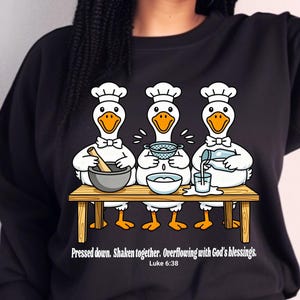 May include: Black sweatshirt featuring a cartoon illustration of three geese wearing chef hats, preparing food at a wooden table. The text "Pressed down. Shaken together. Overflowing with God's blessings." is printed below the image. The word "BLACK" is printed in a black rectangle.