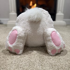 May include: White plush bunny with pink feet and a fluffy tail. The bunny is sitting on a light brown carpet.