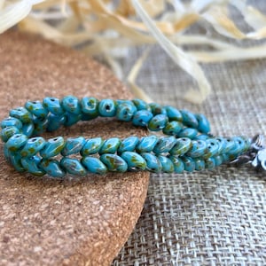 May include: A turquoise beaded bracelet with a silver clasp. The beads are a teardrop shape with a mottled green and brown pattern. The bracelet is displayed on a cork surface.
