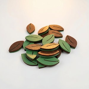 Könnte beinhalten: Eine Ansammlung von Blattausschnitten aus Holz in Braun- und Grüntönen. Die Blätter haben ein detailliertes Aderdesign und sind auf einer weißen Oberfläche verstreut. Bastelbedarf.