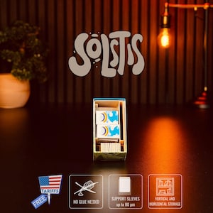 Könnte beinhalten: Eine Holzbox mit Spielkarten mit Regenbogenmuster, darüber das Wort "SOLSTIS". Die Box hat eine blaue Innenseite und wird auf einer dunklen Oberfläche präsentiert. Zusätzliche Symbole weisen auf Funktionen wie "kein Kleber erforderlich" hin.