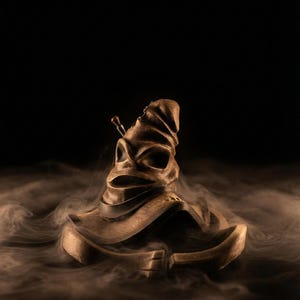 May include: A bronze-colored Sorting Hat, a wizarding hat from the Harry Potter series, sits amidst a cloud of smoke against a black background. The hat has a weathered appearance with folds and creases, and a small, decorative element on top.