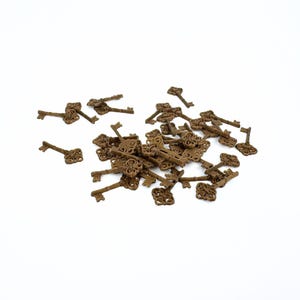 May include: A collection of small, antique-style keys in various orientations. The keys are a bronze colour and have ornate, decorative designs on their heads. They are scattered on a white surface.