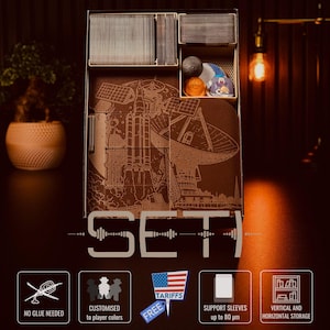 Puede incluir: Un organizador de juegos de madera marrón con compartimentos para cartas y accesorios. La parte delantera presenta un grabado detallado de un diseño de cohete y satélite. La palabra "SET" se muestra de forma destacada. Iconos adicionales.