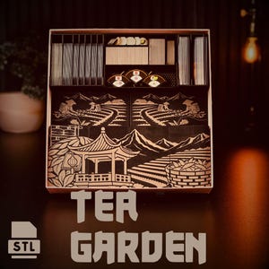 May include: A wooden game box with compartments for game pieces. The box features a black and white illustration of a tea garden with the text "TEA GARDEN" and the STL logo. The box is designed to organize game components.