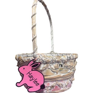 May include: A small, woven Easter basket with a handle. A pink felt bunny tag with the name "Haylee" is attached. The basket is made of natural-colored woven material with a white and pink wash.
