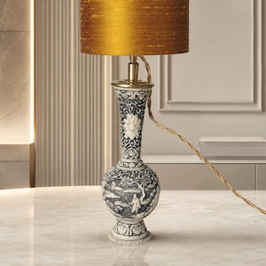 May include: A table lamp featuring a cylindrical mustard-gold fabric lampshade and a tall, slender base made of white and dark gray material. The base is intricately carved with detailed landscape scenes, floral motifs, and a brass-colored neck.