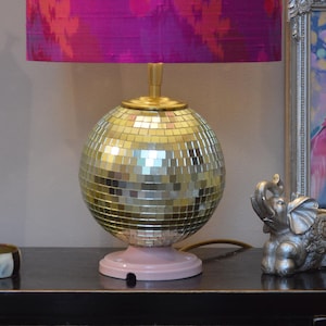 Puede incluir: Una lámpara de mesa decorativa con una base de bola de discoteca espejada y una base rosa. La lámpara tiene un tallo dorado y una pantalla con estampado rosa y morado vibrante. La lámpara está sobre una superficie negra.