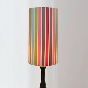 May include: A tall, cylindrical lampshade with a colorful vertical stripe pattern in shades of blue, green, pink, red, gray, and white. The lampshade sits atop a black lamp base. The lamp is a decorative lighting fixture.
