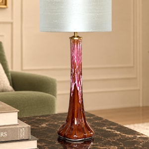 Op de afbeelding: Een tafellamp met een hoge, gegroefde glazen voet met een glanzende bruine onderkant die overgaat in een levendig roze druppelpatroon. De lamp is voorzien van een cilindrische, lichtgrijze stoffen kap en een goudkleurige hals.