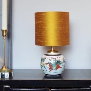 Op de afbeelding: Een decoratieve tafellamp met een gouden zijden lampenkap. De keramische voet heeft een handgeschilderd ontwerp van figuren te paard in rood, groen en blauw. De lamp staat op een zwart oppervlak.