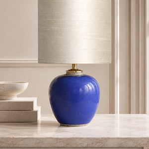 Puede incluir: Una lámpara de mesa con una base de cerámica azul cobalto vibrante con un acabado de esmalte craquelado visible. La lámpara está rematada con una pantalla cilíndrica de tela gris claro e incluye una pieza de cuello dorada que une la base.