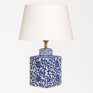 May include: A white lampshade sits atop a blue and white ceramic lamp. The lamp base is square with a floral and vine pattern. The lamp has a brass-colored neck and a white lampshade.