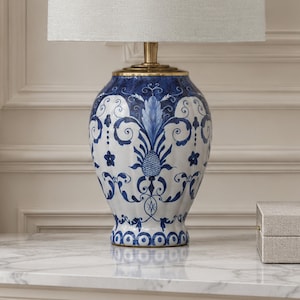 Peut inclure: Une lampe de table avec une base en céramique blanche et bleue ornée d'un motif floral et de volutes détaillé. La lampe possède un col en métal doré et un abat-jour tambour en tissu clair. Le corps en céramique a une forme arrondie.