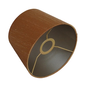 Könnte beinhalten: Ein brauner Lampenschirm mit zylindrischer Form. Der Lampenschirm hat eine strukturierte Außenseite und eine dunkle Innenseite mit einem goldfarbenen Ring und einer Haltestruktur.
