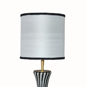 Könnte beinhalten: Weißer Lampenschirm mit schwarzem Besatz, der auf einem schwarz-weiß gestreiften Lampenfuß steht.